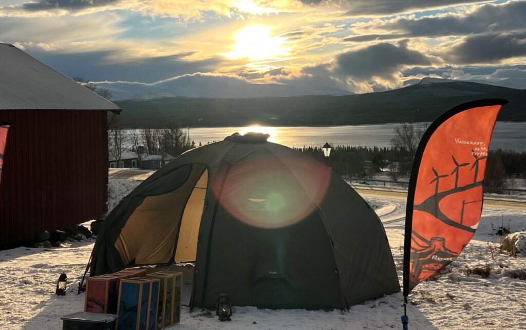 Naturums gröna kupoltält och en orange flagga i förgrunden, i bakgrunden solen och utsikt över Ottsjön och fjällen