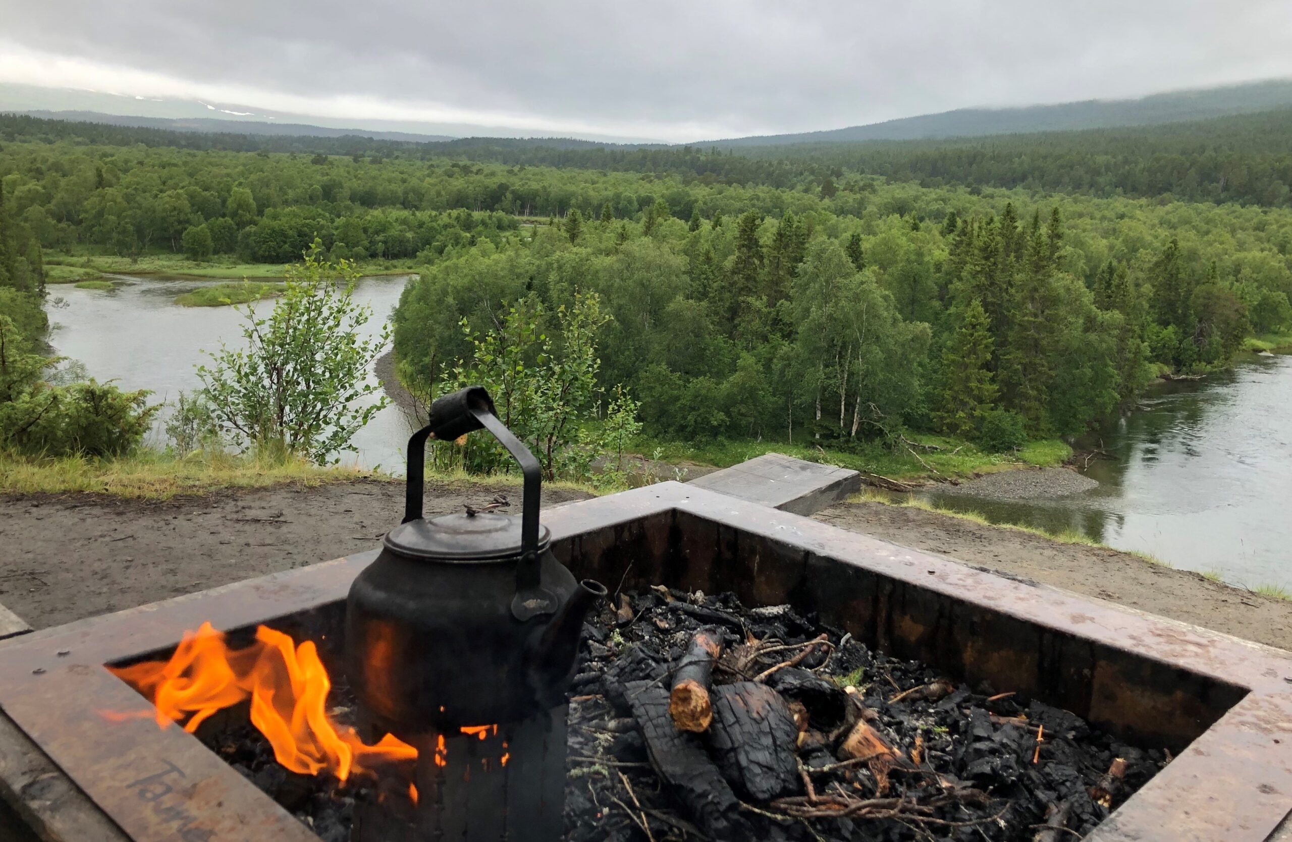 Liten eld och sotig kaffepanna i förgrunden, i bakgrunden utsikt över Vålån och grön skog