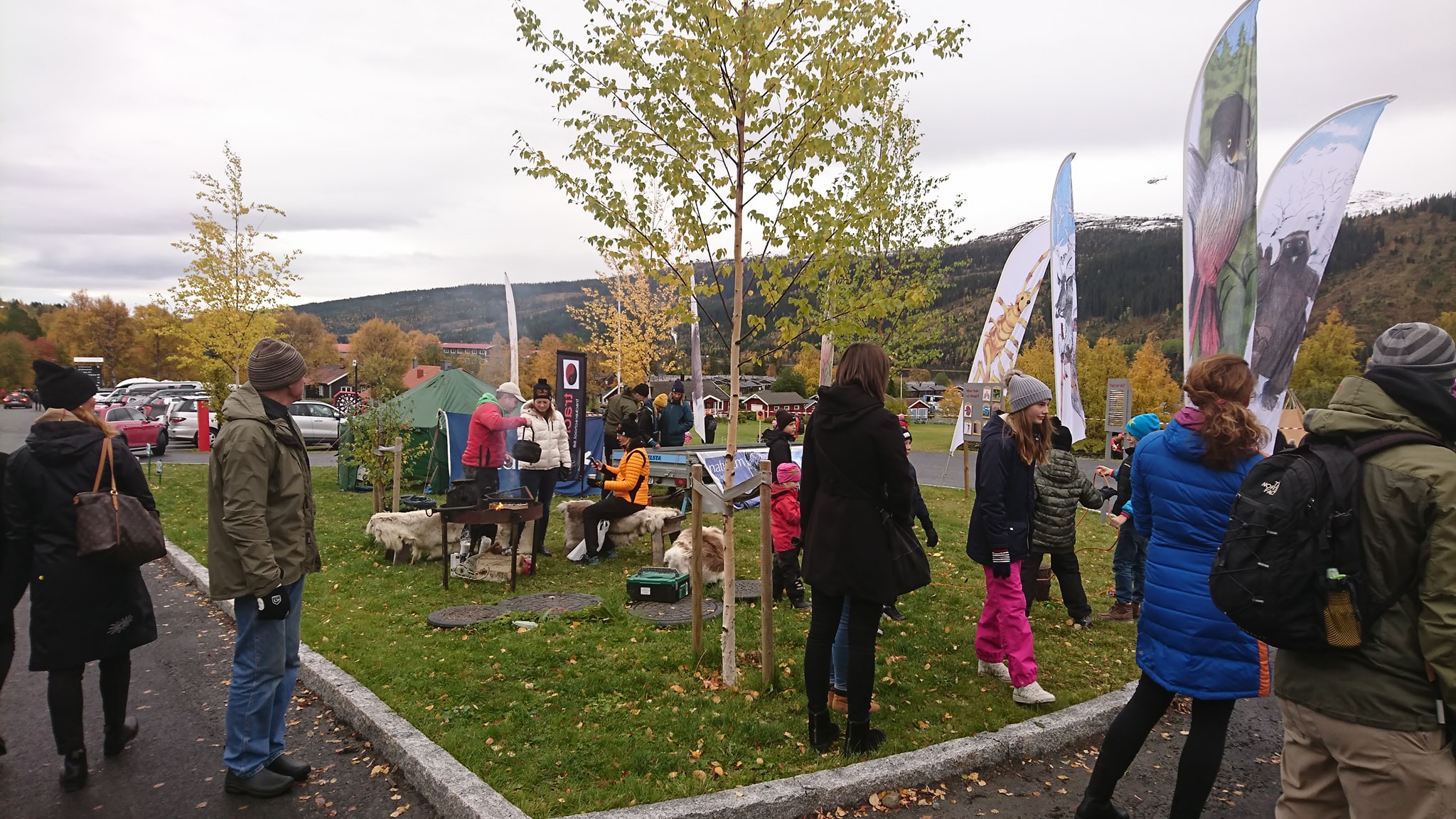 En grupp människor samlade kriing en marknadsaktivitet. I bakgrunden syns björkar och flaggor.