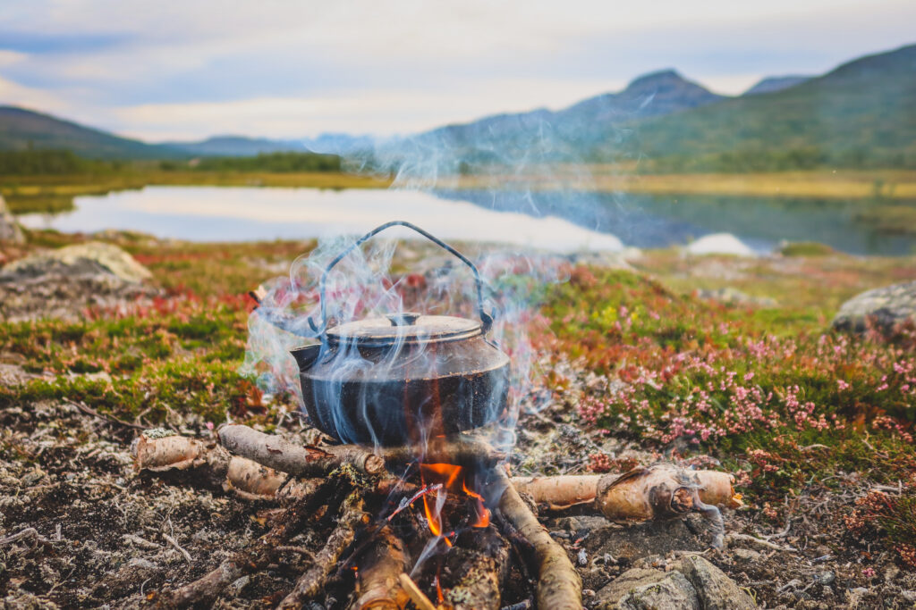 En sotig kafffepanna över öppen eld, med en stilla fjällsjö och höga berg i bakgrunden