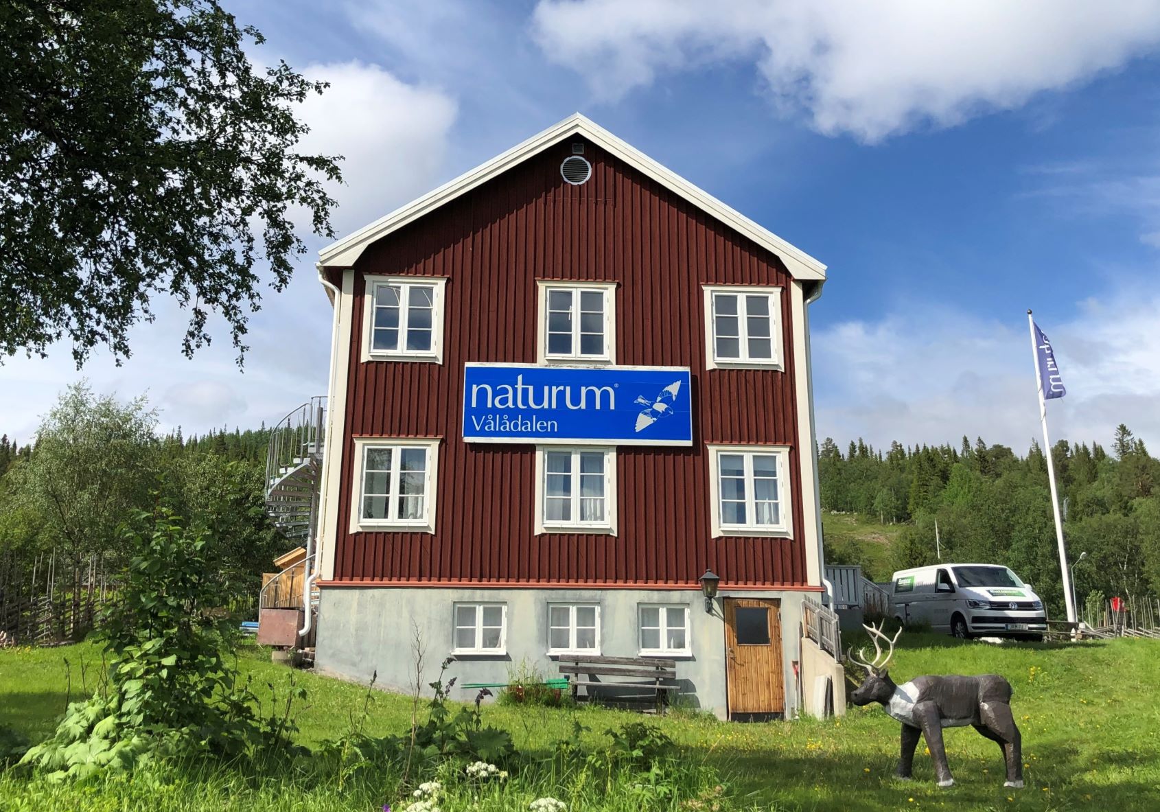 Det röda naturumhuset med vita knutar ch en blå skylt, omgivet av sommargrönska. I förgrunden en av naturums renar i trä.