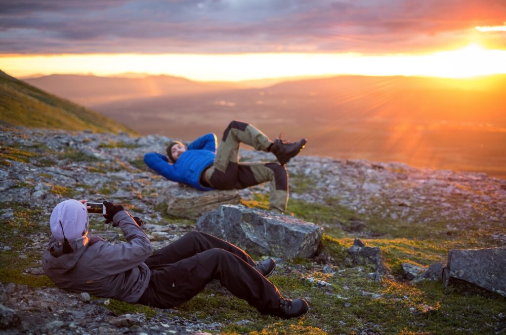 Fjälllandskap med solnedgång och person i förgrunde som ligger på marken och fotograferar annan person i bakgrunden, som också ligger på marken.
