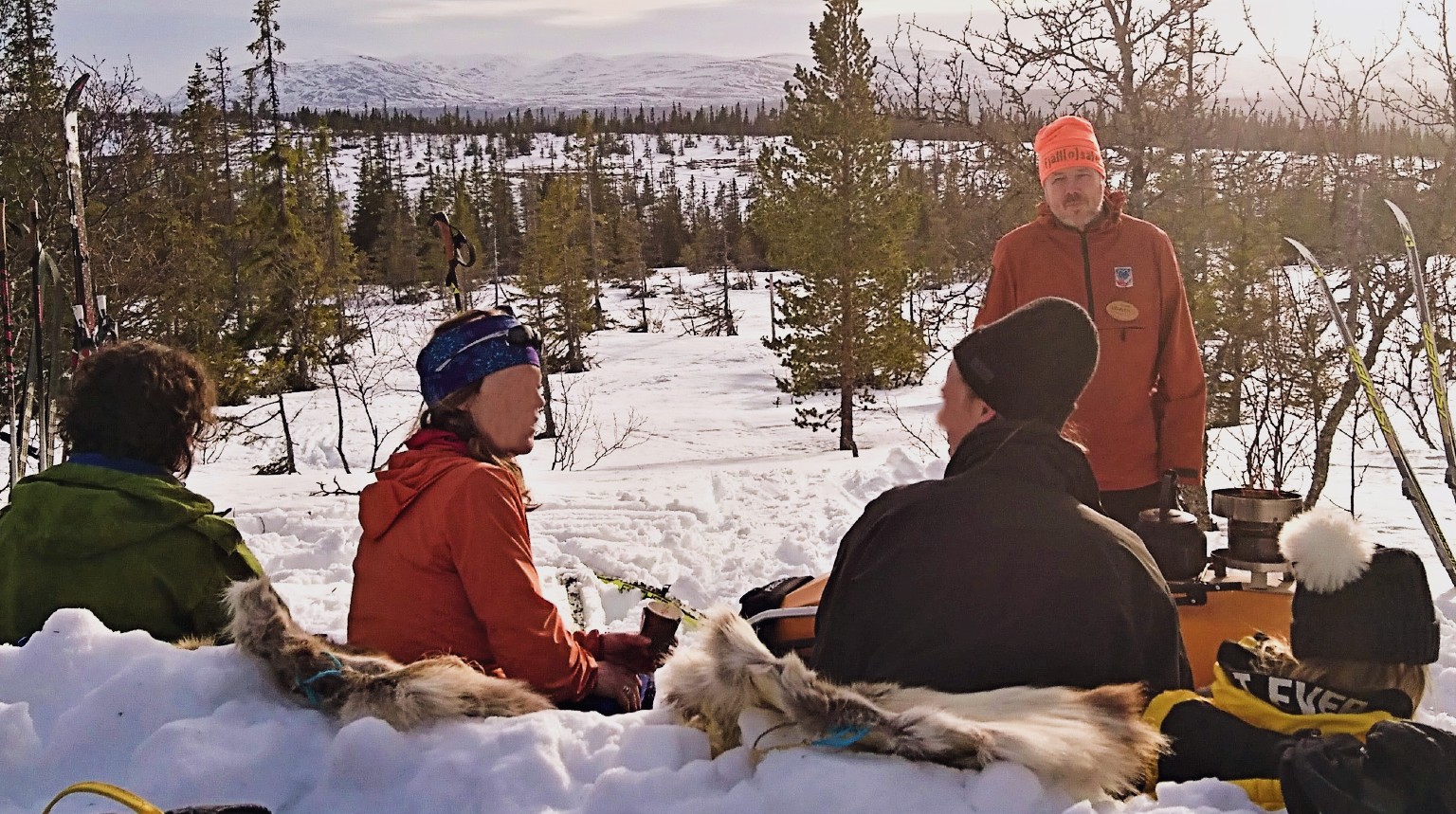 Tre personer sitter på renskinn i en snösoffa med fjällutsikt och pratar med naturums guide