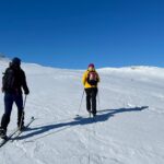 Två personer åker skidor uppför en sluttning på kalfjället. Blå himmel, vit snö.