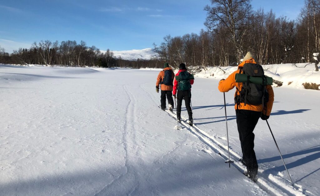 Tre personer åker skidor i soligt vinterlandskap med fjäll i horisonten