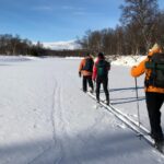 Tre personer åker skidor i soligt vinterlandskap med fjäll i horisonten