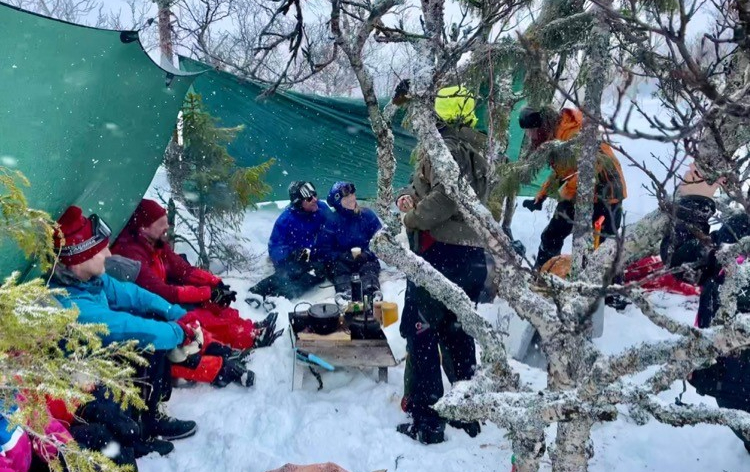 Vinterklädda människor sitter bland fjällbjörkar med uppspänd tarp